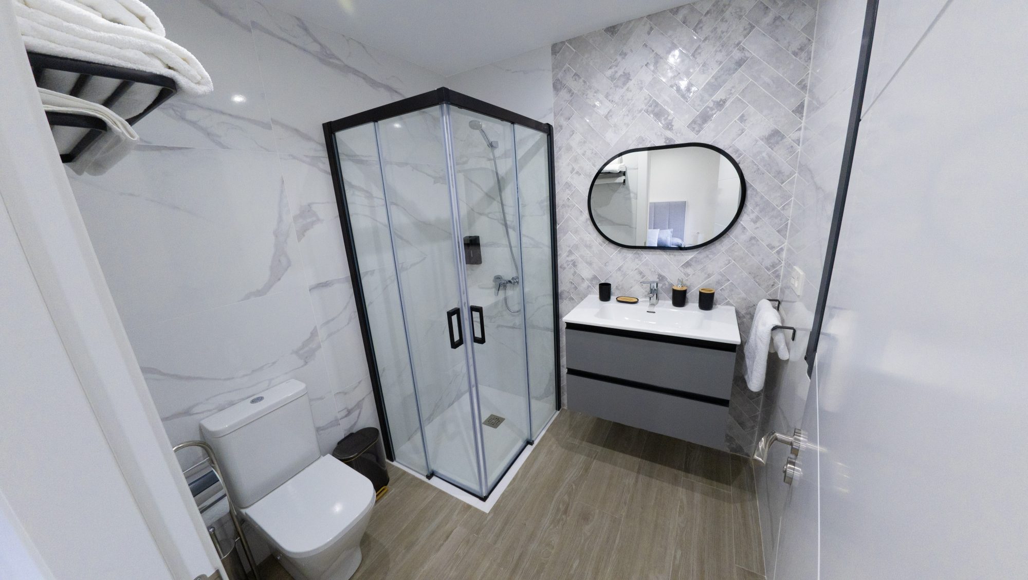 Baño moderno en mármol blanco y herringbone gris con mueble grafito y espejo ovalado.