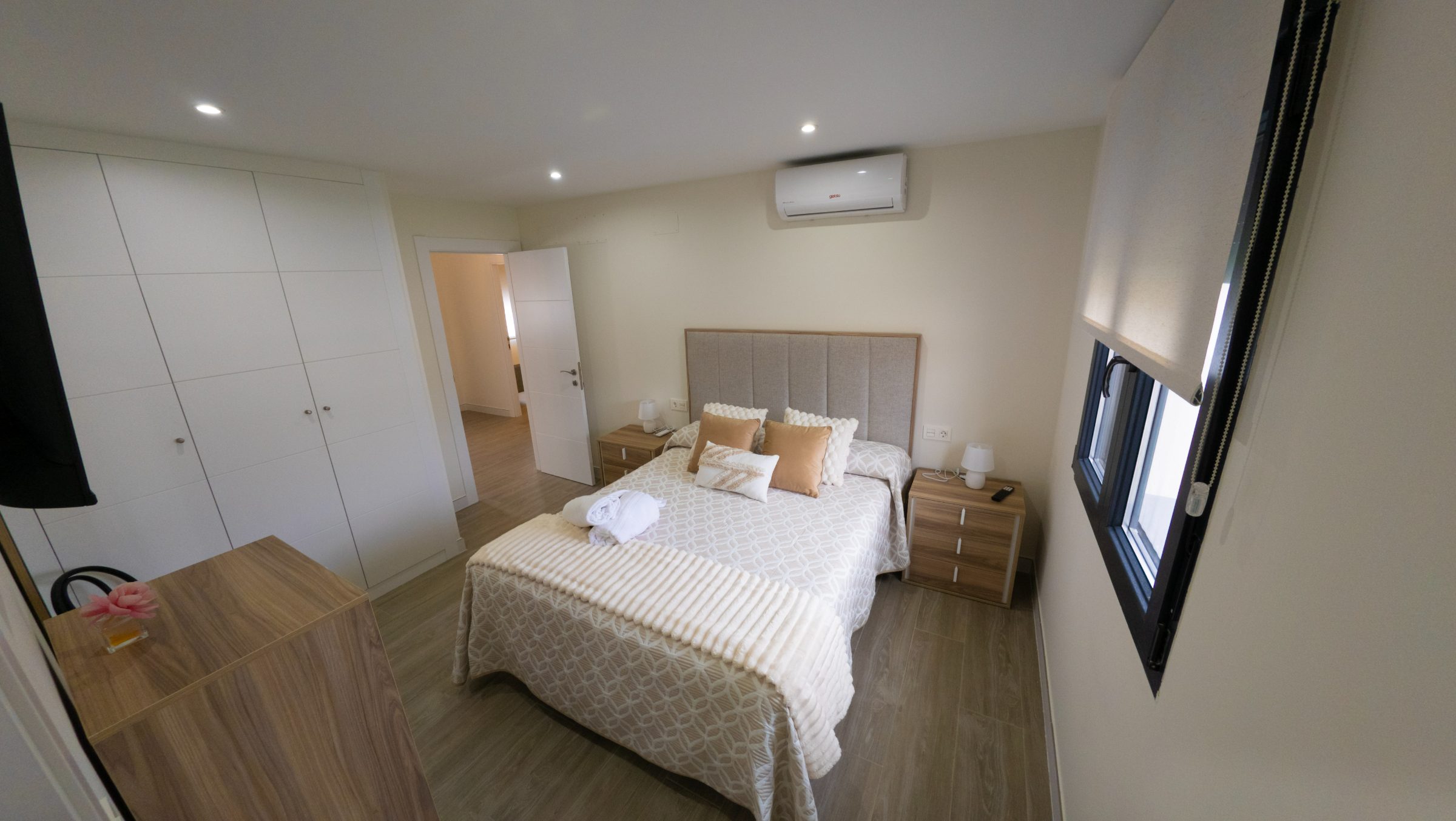 Dormitorio doble en tonos cálidos con cama beige, mesillas de madera y aire acondicionado.